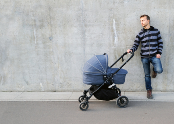 Modern father with newborn stroller standing near gray concrete wall and looking left on copy space. Full length portrait of single parent with baby carriage. Banner.