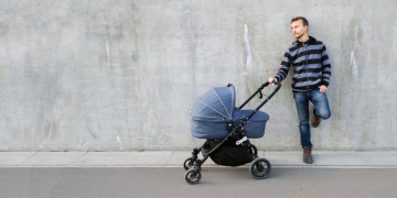 Modern father with newborn stroller standing near gray concrete wall and looking left on copy space. Full length portrait of single parent with baby carriage. Banner.