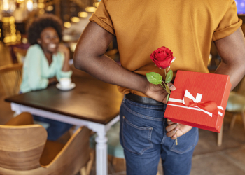 Man hiding red rose behind his back and gift for his smiling girlfriend while sitting in restaurant.
