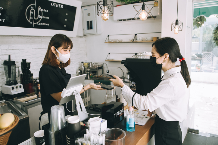 Asian woman cafe owner businesswoman receiving payment from her customer at counter using QR code contactless payment , Two woman wearing a facemask to avoid the spread of coronavirus COVID-19 lifestyle concepts- stock photo