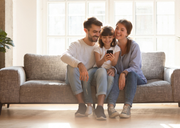 Happy family and kid daughter having fun with smartphone gadget at home, little child girl looking at phone play game using app with mom dad watching funny mobile video, making online call on couch