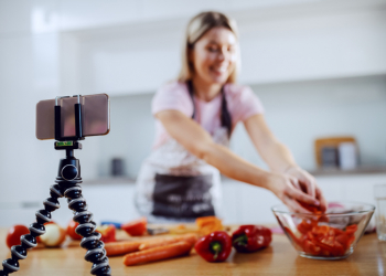 Attractive caucasian blonde woman in apron preparing vegetarian meal. In foreground is smart phone on tripod. Kitchen interior. Selective focus on phone.