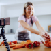 Attractive caucasian blonde woman in apron preparing vegetarian meal. In foreground is smart phone on tripod. Kitchen interior. Selective focus on phone.