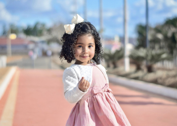 Portrait of a cute little girl hanging out at a park, wearing a pink dress and a white blouse, joyfully enjoying the moment. She is holding some small object and showing it to the camera with a beautiful look on her face.