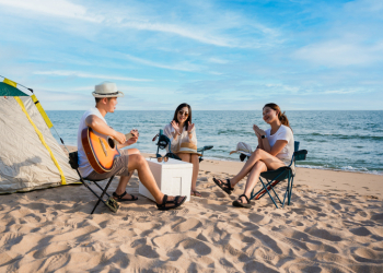 Group of happy Asian friends playing guitar and singing with clap while picnic and camping on the beach in outdoors vacation summer. Young male, female tourist smile fun at party near tent on seaside.