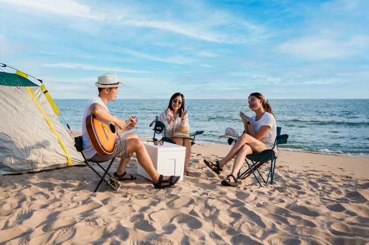 Group of happy Asian friends playing guitar and singing with clap while picnic and camping on the beach in outdoors vacation summer. Young male, female tourist smile fun at party near tent on seaside.