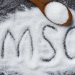 Heap of monosodium glutamate on wooden spoon and dark background with text MSG