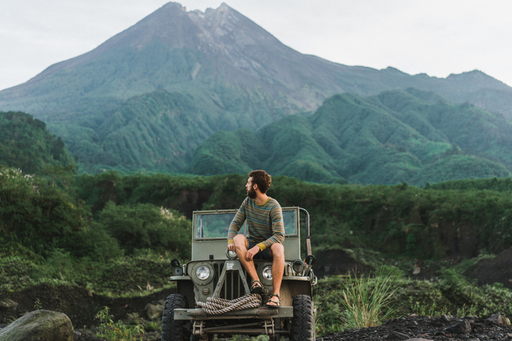 Scenic view of young Caucasian man sitting on old fashioned SUV on the background of Merapi volcano