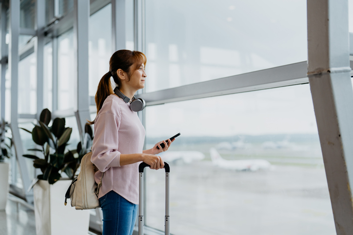 Image of an Asian Chinese woman tourist holding smartphone in airport terminal and looking at airport runway