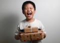 Portrait of a happy Asian boy holding a stack of Christmas gifts against a grey wall.