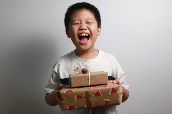 Portrait of a happy Asian boy holding a stack of Christmas gifts against a grey wall.